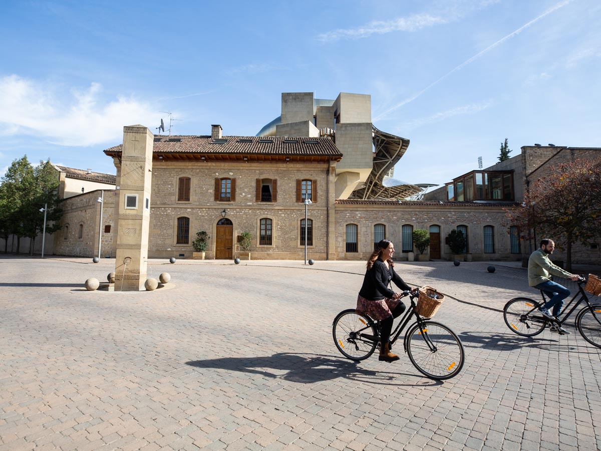 Dos turistas recorren en bici la plaza del reloj en la ciudad del vino de Marqués de Riscal. 